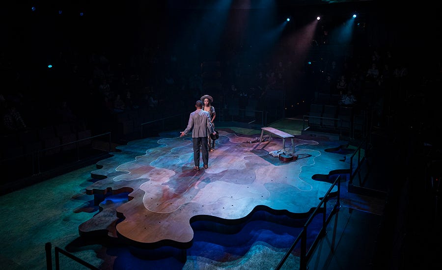 Leroy, a young Black man, has his back to the camera, wearing black pinstripe pants and a gray blazer. He is facing Lucy, a young Black woman wearing a floral dress and a straw hat. They stand on an abstract set, bathed in green and blue light.