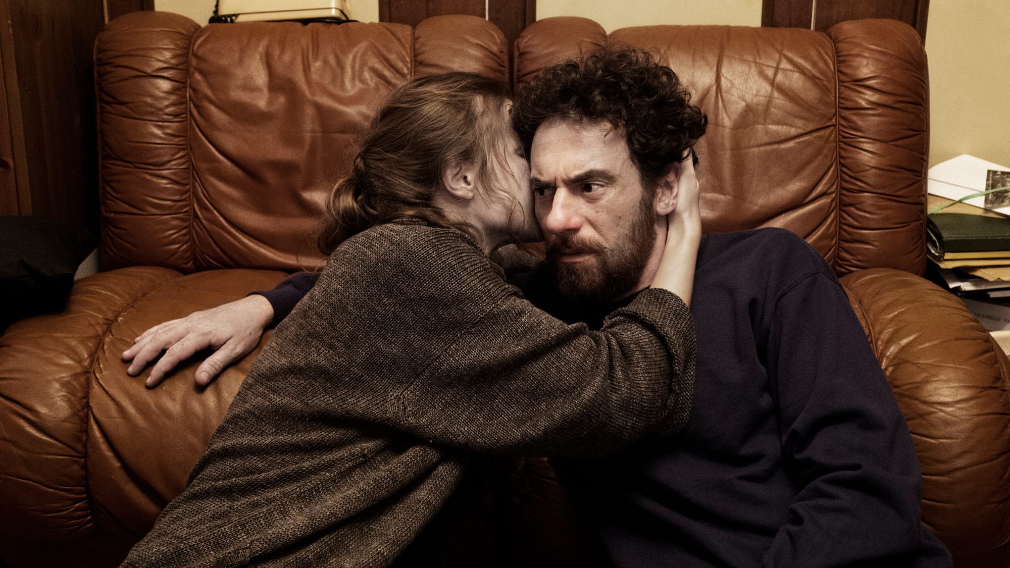 A woman in her 20s, wearing a brown sweater, whispers in the ear of a man in his mid-30s wearing a maroon sweater. Both lean on a brown leather couch.