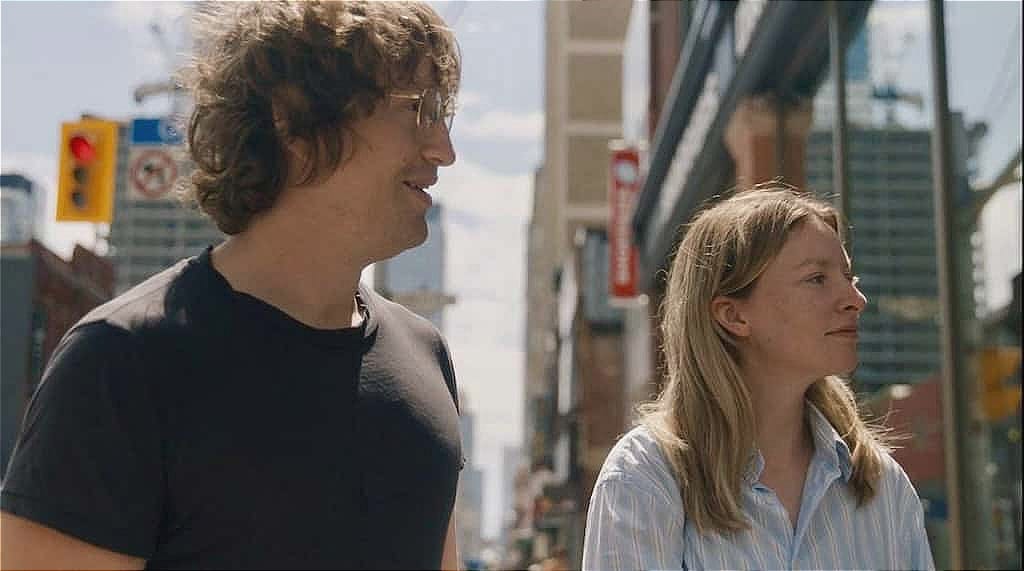 A white man in his 30s, with coke-bottle glasses and wearing a white t-shirt, stands next to a white woman in her mid-30s wearing a striped button-down.