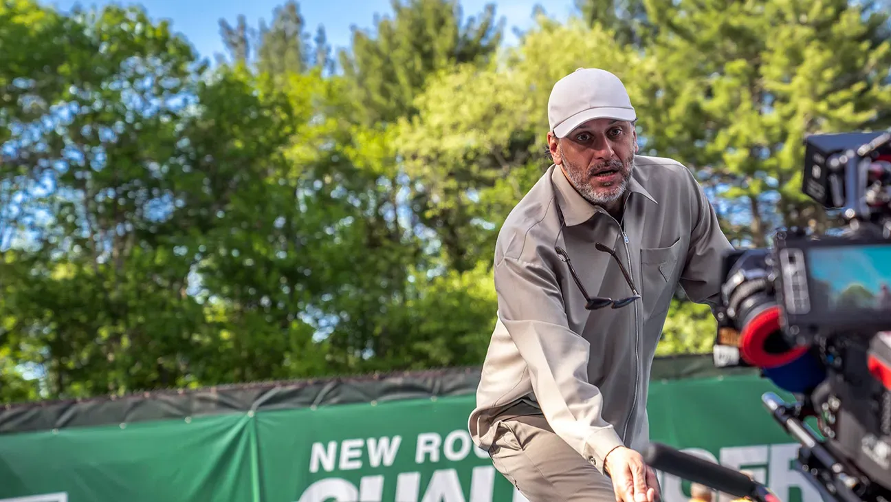 A man in his 50s wearing a gray shirt and ball cap with sunglasses on a chain positions a camera.