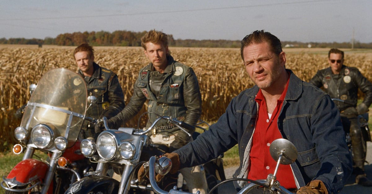 Four white men ranging in age from their 20s to their 50s, all wearing denim jackets, sit on motorcycles.