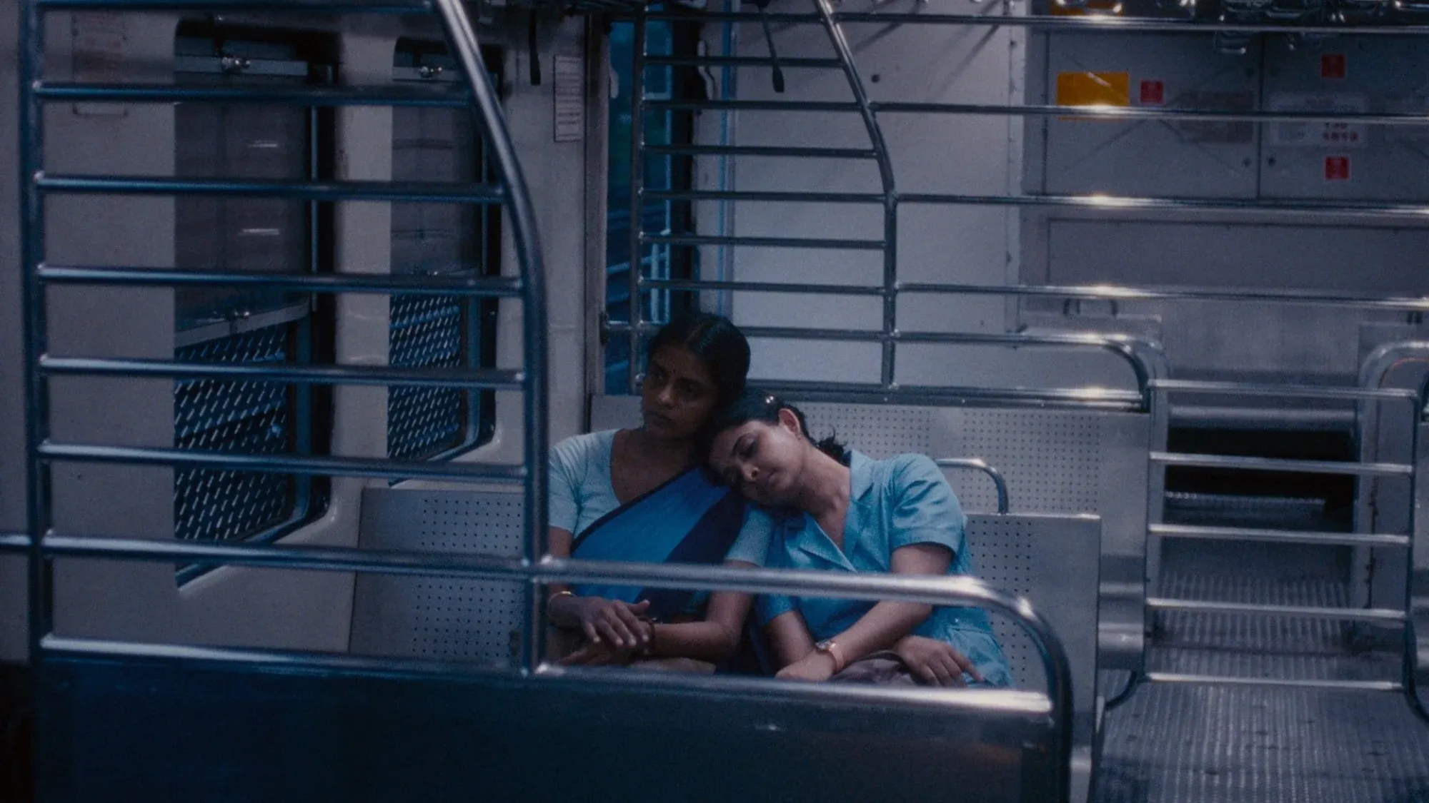 Two Indian women in their 30s share a bench in a rail car. Both wear light blue scrubs. One has her head on the other's shoulder.