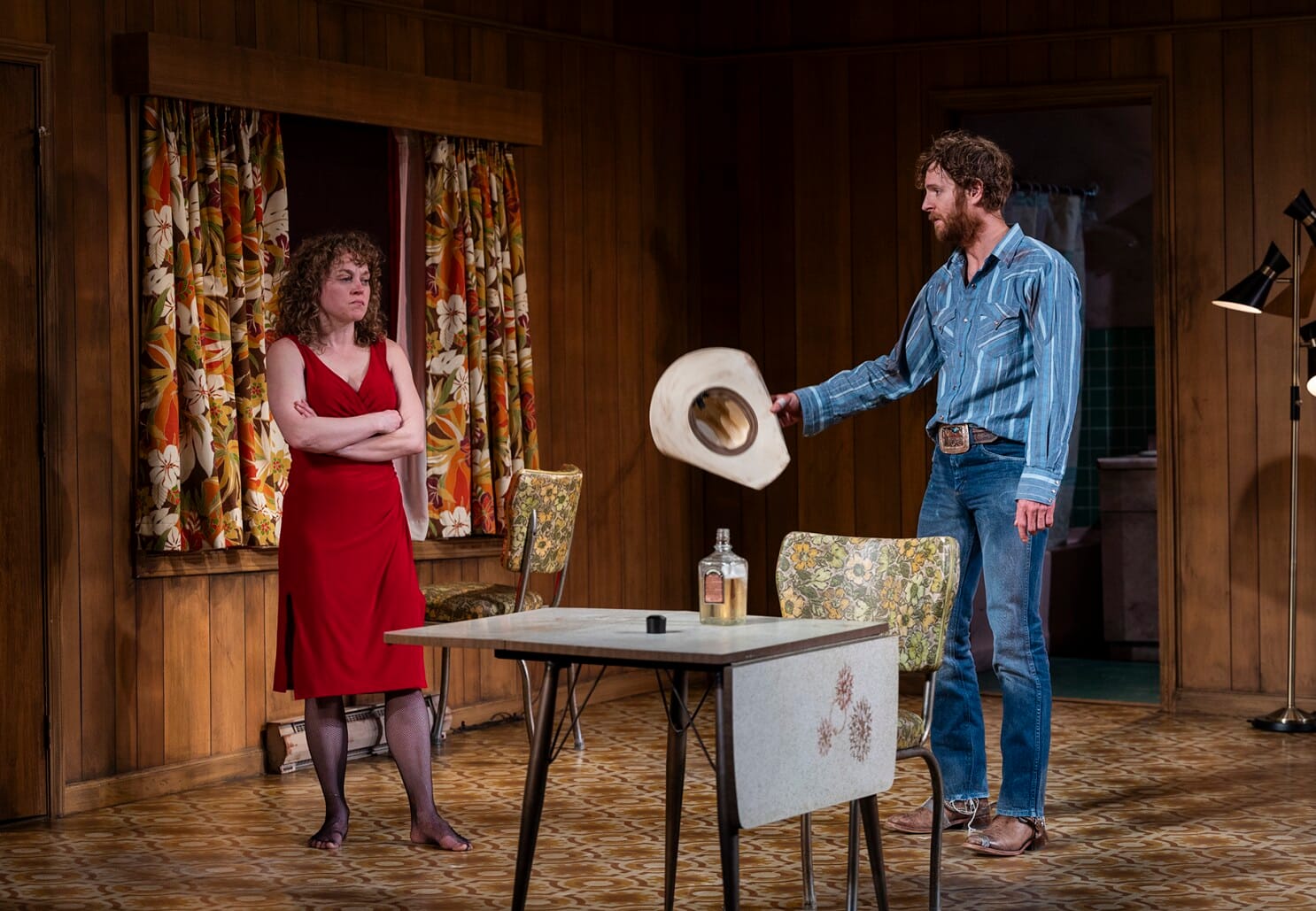 A white woman in her 30s, wearing a red dress and sheer black tights, stands across from a white man in his 30s wearing Western wear. He gesticulates with his cowboy hat. Between them is a bottle of tequila.