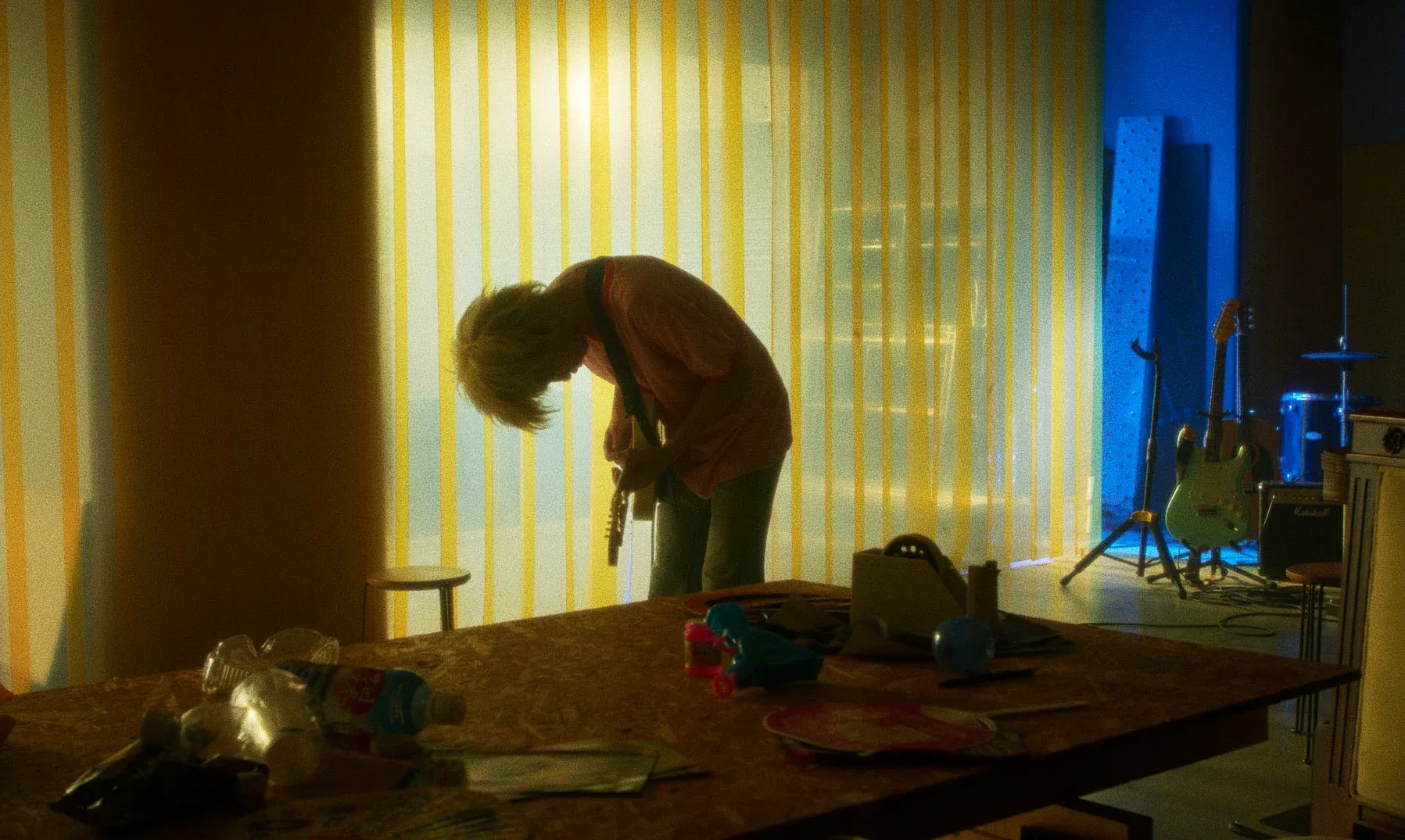 A young Japanese man is hunched over his guitar in a dimly-lit room.