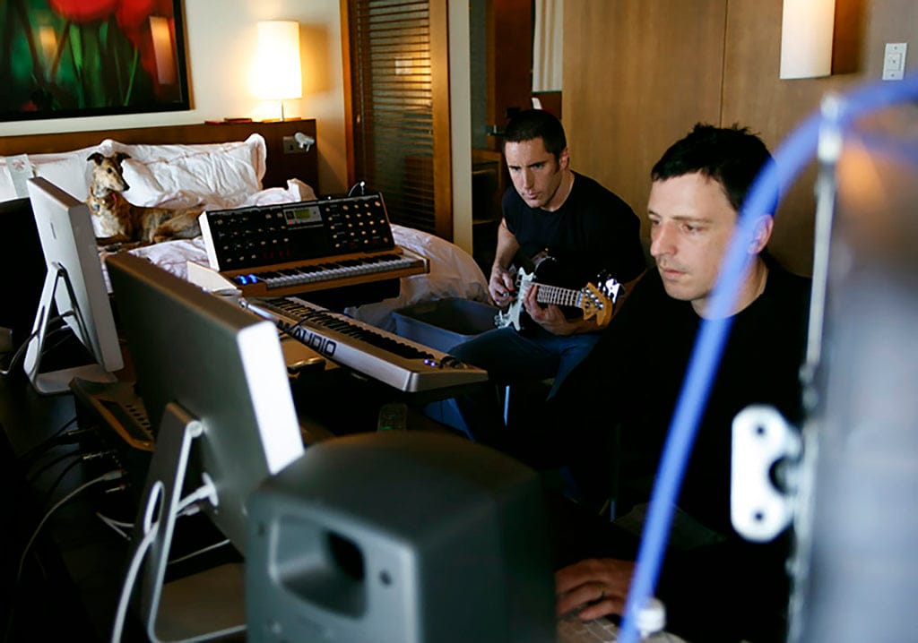 A white man in his 40s plays bass while another white man in his 40s works on a computer. Both wear all black. A greyhound looks on.