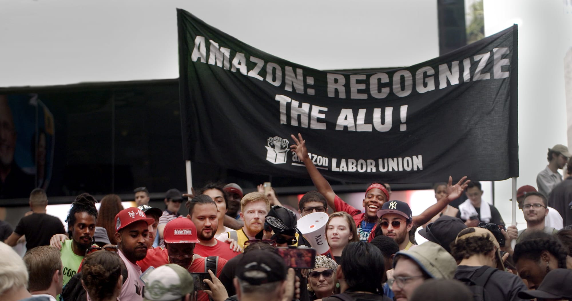 A crowd of workers stand in front of a black banner that reads: "Amazon: Recognize the ALU! Amazon Labor Union".