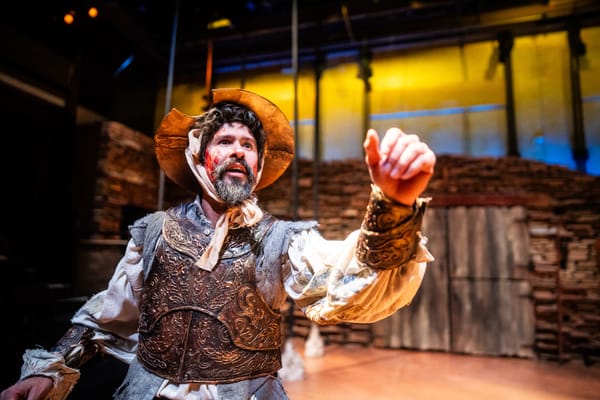 A man in his 30s wears armor, a ratty linen shirt, and a golden bowl on his head. He has stage blood on his face as well.