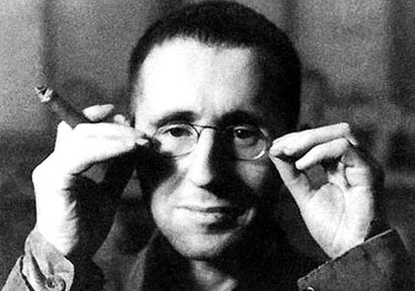 Bertolt Brecht, a white, middle-aged man adjusts his glasses, with a cigar between the fingers on his right hand.