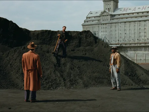 A middle-aged white man stands on a mound of dirt, flanked by a white man in a long brown coat and a black man wearing jeans.